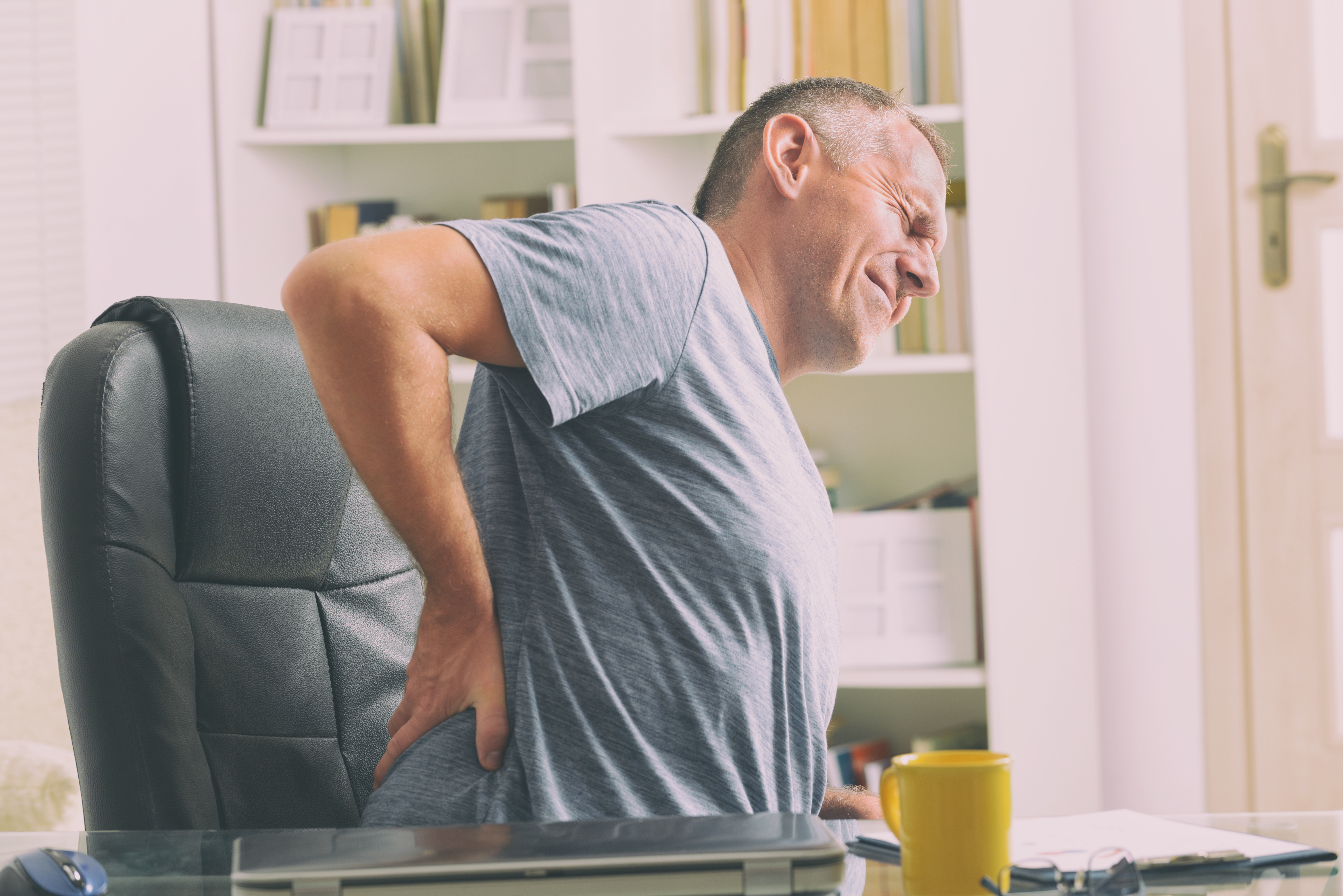 Man in home office suffering from low back pain sitting at the desk with notebook, papers and other objects